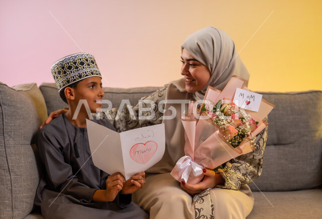 المفاجآت وهدايا العيد, بورتريه لولد عربي خليجي عماني يرتدي الدشداشة والكمه يقوم بإعطاء أمه باقة من الورد وكرت معايدة, مفهوم الإهداء وتقدير الوالدين, عائلة عمانية تحتفل بالعيد المبارك, أجواء لمة الأهل في المناسبات والاحتفالات السعيدة, خلفية ملونة