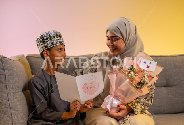تقديم الزهور والهدايا للأم, بورتريه لولد عربي خليجي عماني يرتدي الدشداشة والكمه يقوم بإعطاء أمه باقة من الورد, مفهوم الإهداء وتقدير الوالدين, عائلة عمانية تحتفل بالعيد المبارك, أجواء لمة الأهل في المناسبات والاحتفالات السعيدة, خلفية ملونة