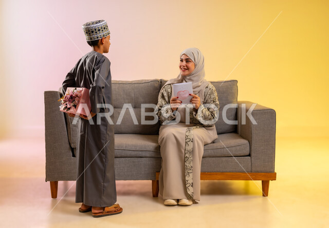إعطاء الأم هدية العيد, بورتريه لولد عربي خليجي عماني يقوم بإعطاء أمه علبة مغلفة بتنسيق جميل, مفهوم الإهداء وتقدير الوالدين, عائلة عمانية تحتفل بالعيد المبارك, أجواء لمة الأهل في المناسبات والاحتفالات السعيدة, إيماءات السعادة والمفاجأة بالهديه, خلفية ملونة