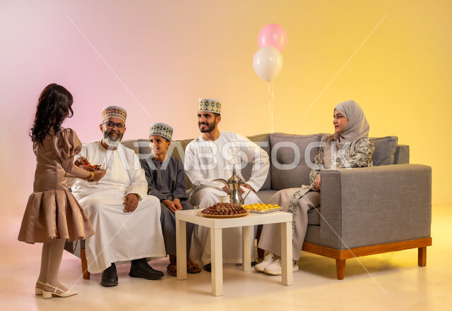 تبادل التهنئات والمعايدات بين أفراد الأسرة بإيماءات الفرح والسعادة, أجواء العيد بين الأهل, بورتريه لعائلة عربية خليجية عمانية تحتفل بالعيد السعيد, فتاة تقوم بتقديم الحلويات والضيافة الشعبية, طقوس الأعياد والمناسبات الاسلامية في سلطنة عمان, خلفية ملونة