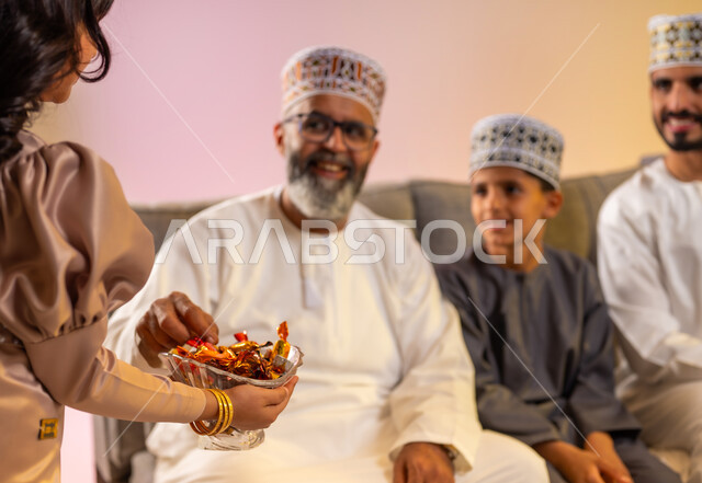 Eid sweets for visitors and guests, a girl providing popular hospitality, rituals of Islamic holidays and occasions in the Sultanate of Oman, exchange of congratulations and greetings between family members with gestures of joy and happiness, portrait of an Omani Gulf Arab family celebrating the happy Eid, colorful background