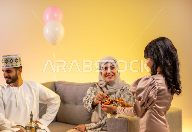 ضيافة العيد للزوار والضيوف, فتاة تقوم بتقديم الحلويات الشعبية, طقوس الأعياد والمناسبات الاسلامية في سلطنة عمان, تبادل التهنئات والمعايدات بين أفراد الأسرة بإيماءات الفرح والسعادة,  بورتريه لعائلة عربية خليجية عمانية تحتفل بالعيد السعيد, خلفية ملونة