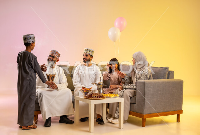 طقوس الاحتفالات في صباح العيد، تجمع العائلة والجلوس برفقة بعضها البعض بسعادة, ولد يرتدي الدشداشة والكمه يقوم بالتبخير واستخدام عطور العود الفاخرة لاستقبال العيد المبارك, بورتريه لعائلة عربية خليجية عمانية تحتفل بالمناسبات الاسلامية , خلفية ملونة