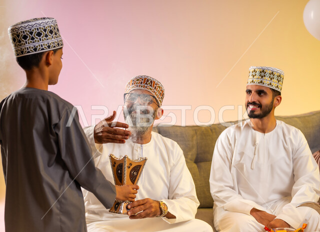تقاليد وطقوس الاحتفالات في صباح العيد، تجمع العائلة والجلوس برفقة بعضها البعض بسعادة, بورتريه مقرب لولد يرتدي الدشداشة والكمه يقوم بالتبخير واستخدام عطور العود الفاخرة لاستقبال العيد المبارك, عائلة عربية خليجية عمانية تحتفل بالمناسبات الاسلامية , خلفية ملونة