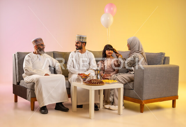 Sitting in the living room in a warm family atmosphere, wearing traditional Omani clothes, family gathering to receive the blessed Eid, showing feelings of tenderness, love and affection, portrait of an Omani Gulf Arab family celebrating Islamic occasions in the company of each other, colorful background