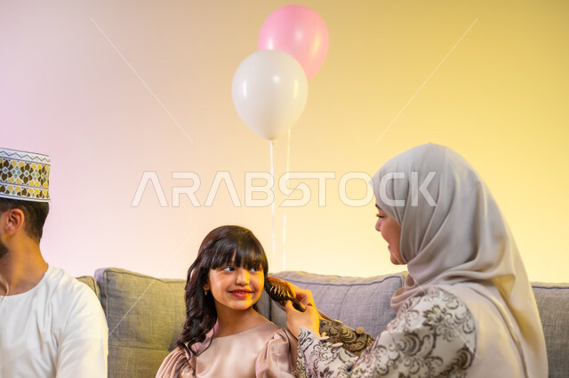 الاحتفالات المبهجة في صباح العيد، تجمع العائلة والجلوس برفقة بعضها البعض بسعادة, بورتريه مقرب لأم محجبة تقوم بتسريح شعر ابنتها الصغيرة لاستقبال العيد المبارك, عائلة عربية خليجية عمانية تحتفل بالمناسبات الاسلامية , ارتداء الملابس العمانية التقليدية, خلفية ملونة