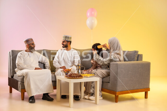الطقوس المبهجة في صباح العيد، تجمع العائلة والجلوس برفقة بعضها البعض بسعادة, بورتريه لعائلة عربية خليجية عمانية تحتفل بالمناسبات الاسلامية, ارتداء الملابس العمانية التقليدية, أم محجبة تقوم بتسريح شعر ابنتها الصغيرة لاستقبال العيد المبارك, خلفية ملونة