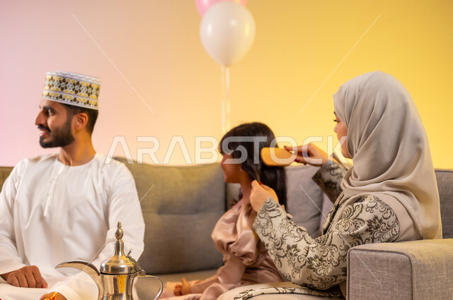 الاهتمام بالأناقة والمظهر الخارجي، طقوس تجمع العائلة والجلوس برفقة بعضها بسعادة, بورتريه مقرب لأم محجبة تقوم بتسريح شعر ابنتها الصغيرة لاستقبال العيد المبارك, عائلة عربية خليجية عمانية تحتفل بالمناسبات الاسلامية, ارتداء الملابس العمانية التقليدية, خلفية ملونة