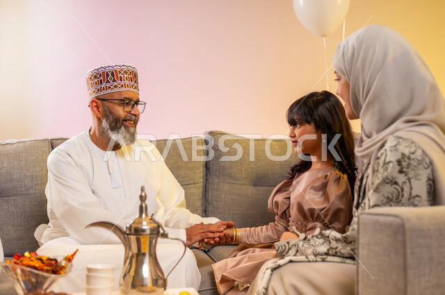 اجتماع الجد مع حفيدته وإظهار مشاعر الحنان والمحبة, الابتهاج بقدوم العيد المبارك, الجلوس في غرفة المعيشة في جو عائلي دافئ، بورتريه لعائلة عربية خليجية عمانية تحتفل بالمناسبات الاسلامية برفقة بعضها البعض, ارتداء الثياب الجديدة الجميلة, خلفية ملونة