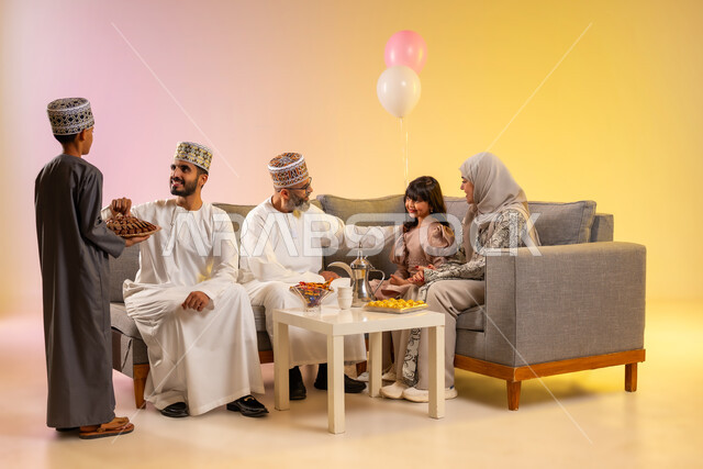 مساعدة الأبناء في تقديم الضيافة، ولد يرتدي الدشداشة والكمه يقوم بتوزيع الحلويات الشعبية، المحبة والمودة بين أفراد الأسرة، التهنئات وطقوس المناسبات الاسلامية، الجلوس في غرفة المعيشة في جو عائلي، بورتريه لعائلة عربية خليجية عمانية تحتفل بقدوم العيد, خلفية ملونة
