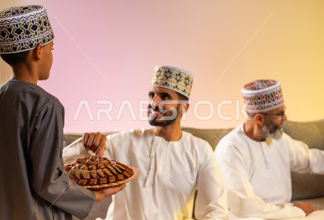 قيام الأبناء بتوزيع الضيافة، ولد يرتدي الدشداشة والكمه يقوم بتقديم الحلويات الشعبية، المحبة والمودة بين أفراد الأسرة، التهنئات وطقوس المناسبات الاسلامية، الجلوس في غرفة المعيشة في جو عائلي، بورتريه لعائلة عربية خليجية عمانية تحتفل بقدوم العيد, خلفية ملونة