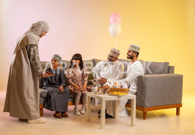 تقديم الضيافة العربية والحلويات الشعبية، المعايدات والتهنئات وطقوس المناسبات الاسلامية، الزيارات والجلوس في غرفة المعيشة في جو عائلي، بورتريه لعائلة عربية خليجية عمانية ترتدي الأزياء التقليدية احتفالاً بقدوم العيد, مفهوم المحبة والمودة، خلفية ملونة