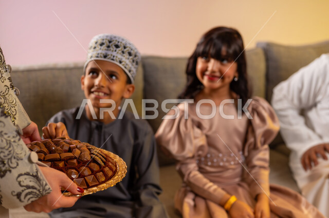 سعادة الأولاد بتقديم الضيافة والحلويات لهم، بورتريه مقرب لأخ وأخته عربيين خليجيين عمانيين يرتديان الملابس الجديدة ويحتفلان بقدوم العيد، الجلوس في غرفة المعيشة بجو من المحبة والمودة بين أفراد الأسرة، الزيارات العائلية والتهنئات وتبادل المعايدات، خلفية ملونة