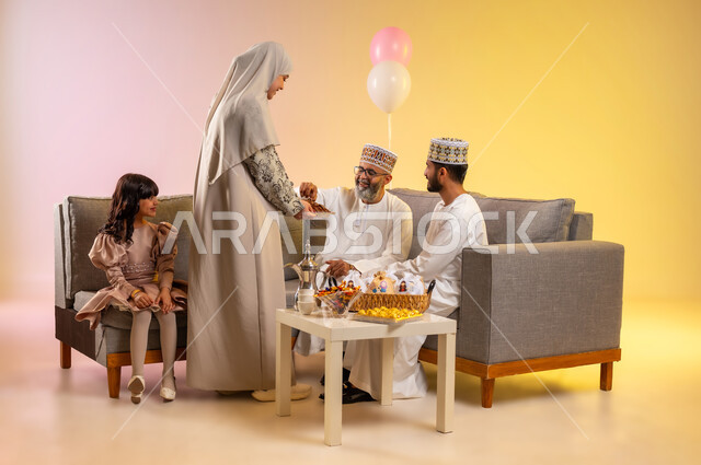 المعايدات والتهنئات وطقوس المناسبات الاسلامية، الجلوس في غرفة المعيشة في جو عائلي والزيارات، بورتريه لعائلة عربية خليجية عمانية ترتدي الأزياء التقليدية احتفالاً بقدوم العيد, تقديم الضيافة العربية والحلويات الشعبية، مفهوم المحبة والمودة، خلفية ملونة