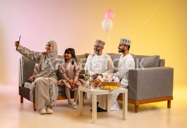 Using a mobile phone to take souvenir photos, sitting in the living room in a family atmosphere and exchanging greetings, a portrait of an Omani Gulf Arab family wearing traditional costumes to celebrate the coming of Eid, providing Arab hospitality and popular sweets, the concept of love and affection, a colorful background.