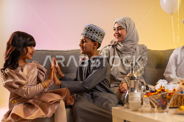 تبادل التهنئات والزيارات العائلية، بورتريه لعائلة عربية خليجية عمانية تجلس في غرفة المعيشة وتحتفل بالعيد في جو عائلي سعيد, تقديم الضيافة العربية والحلويات الشعبية، أخ وأخته يقومان باللعب والمرح والاستمتاع بقضاء الوقت، مفهوم الحنان والمحبة والمودة، خلفية ملونة