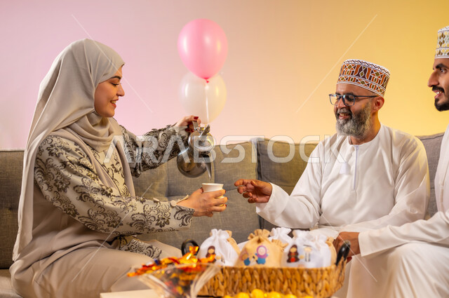 تقديم القهوة العربية والحلويات الشعبية، الاحتفال بالأعياد الإسلامية والزيارات العائلية، بورتريه لعائلة عربية خليجية عمانية تجلس في غرفة المعيشة وتقوم بتبادل المعايدات, إظهار مشاعر الفرح والسعادة بقدوم العيد، مفهوم المحبة والمودة بين أفراد الأسرة، خلفية ملونة