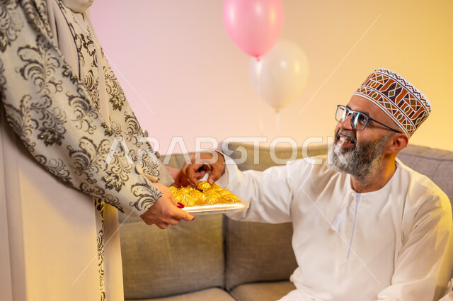 Celebrations of happy occasions in the Sultanate of Oman, a veiled woman serving popular sweets on holidays, a close-up portrait of an Omani Gulf Arab family celebrating the blessed Eid, wearing traditional Omani costumes, the concept of generosity and providing Arab hospitality, a colorful background.