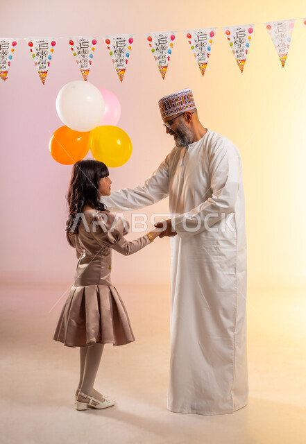 الإمساك بالبالونات الملونة وتعليق حبال الزينة، بورتريه لعائلة عربية خليجية عمانية تحتفل بالأعياد الإسلامية, جد يقوم بالمرح والاحتفال مع حفيدته الصغيرة، إظهار مشاعر الفرح والسعادة بقدوم العيد، مفهوم الحنان والمحبة والمودة،الجسم بالطول الكامل، خلفية ملونة