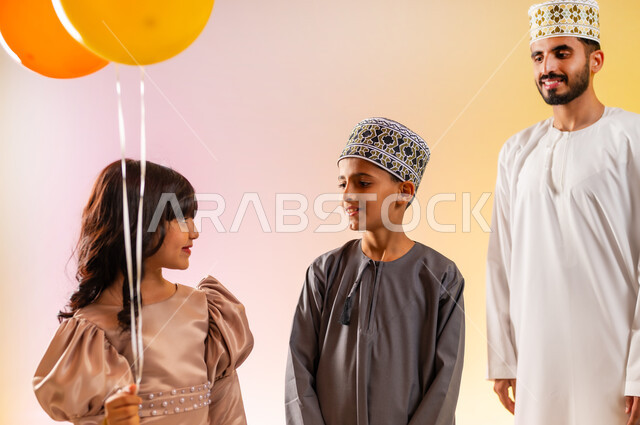 إظهار مشاعر الفرح والسعادة بالمناسبات والأعياد الاسلامية، بورتريه مقرب لعائلة عربية خليجية عمانية تحتفل بالعيد المبارك، فتاة تقوم بالامساك بالبالونات الملونة، ارتداء الأزياء العمانية التقليدية، الاحتفال برفقة أفراد الأسرة وزيادة روابط المحبة، خلفية ملونة