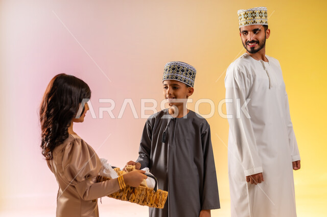 تقديم الحلويات الشعبية في الأعياد، طقوس الاحتفالات والمناسبات السعيدة في سلطنة عمان، بورتريه مقرب لفتاة تقوم بتوزيع الضيافة العربية، عائلة عربية خليجية عمانية تحتفل بالعيد المبارك، ارتداء الأزياء العمانية التقليدية، إظهار مشاعر الفرح والسعادة، خلفية ملونة