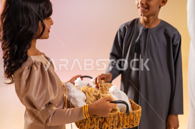 أجواء الاحتفالات والمناسبات السعيدة في سلطنة عمان، توزيع الحلويات الشعبية في الأعياد، بورتريه مقرب لفتاة تقوم بتقديم الضيافة لأخيها، عائلة عربية خليجية عمانية تحتفل بالعيد المبارك، ارتداء الأزياء العمانية التقليدية، إظهار مشاعر الفرح والسعادة، خلفية ملونة