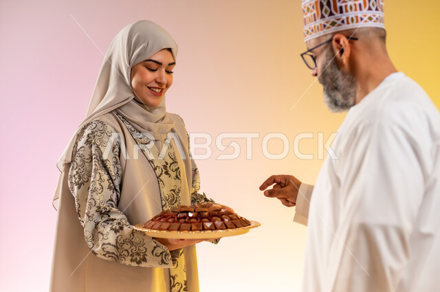 Popular Arab holiday hospitality in the Sultanate of Oman, family affection and love, a portrait of an Omani Gulf Arab family celebrating Islamic holidays, a veiled woman wearing an abaya serving traditional sweets, rituals of celebrations and happy occasions, a colorful background