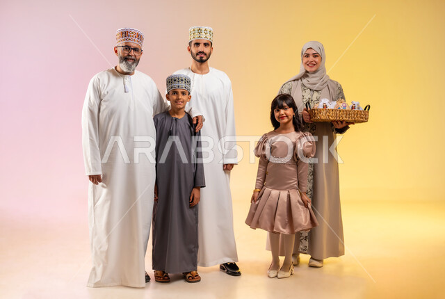 ارتداء الثياب الجديدة وأجواء البهجة في العيد, بورتريه لعائلة عربية خليجية عمانية تحتفل بالأعياد الاسلامية, الوقوف والنظر للكاميرا بإيماءات السعادة والفرح، تقديم الحلويات التقليدية والمأكولات الشهية, إظهار مشاعر المودة والمحبة العائلية, خلفية ملونة