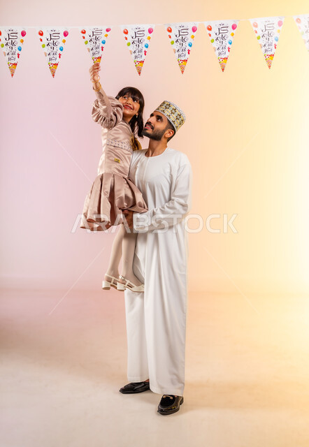 Tenderness of fatherhood and feelings of love and affection, a portrait of an Arab Gulf Omani family celebrating Islamic holidays, a father carrying his young daughter, hanging decorative ropes, showing feelings of joy and happiness, rituals of celebrations and happy occasions, full-length body, colorful background.