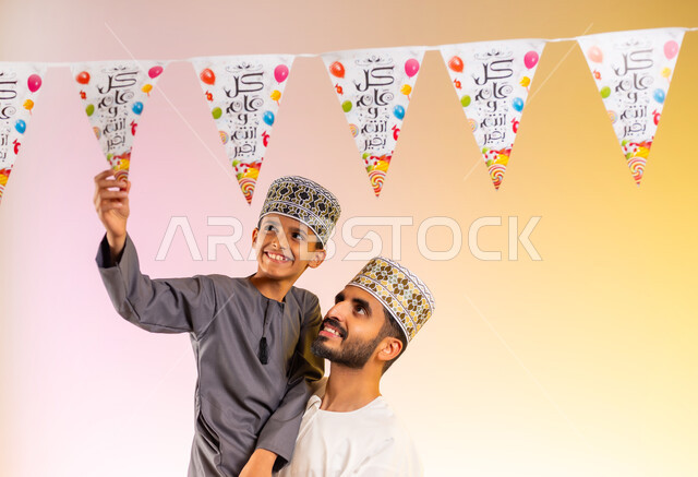 مفهوم الأسرة المتماسكة وحنان الأبوة، إظهار مشاعر المودة والفرح والسعادة, بورتريه لعائلة عربية خليجية عمانية تحتفل بالأعياد الإسلامية, أب يحمل ابنه يرتديان الدشداشة والكمه يقومان بتعليق حبال الزينة، طقوس الاحتفالات والمناسبات السعيدة، خلفية ملونة