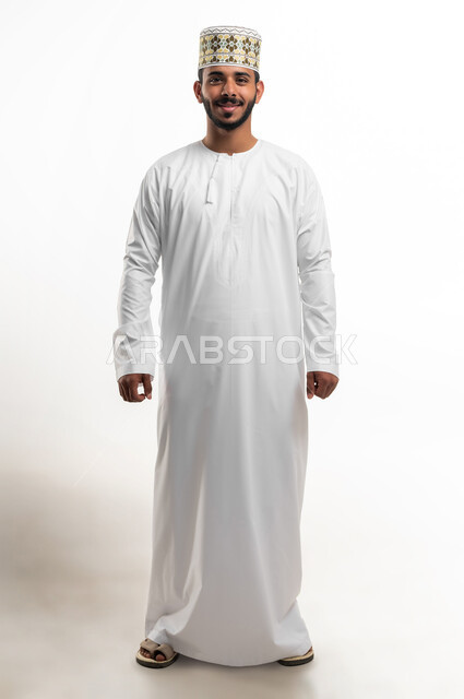 الاهتمام بالأناقة والمظهر الخارجي, مفهوم الرجولة والثقة بالنفس، التقاط الصور الشخصية للوثائق الرسمية، بورتريه لشاب عربي خليجي عماني مبتسم يرتدي الدشداشة و الكمه ينظر الى الكاميرا بإيماءات السرور والسعادة، صورة الجسم بالطول الكامل, خلفية بيضاء