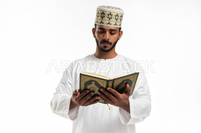 التقرب الى الله في شهر رمضان، الاجتهاد في العبادة، تلاوة وقراءة القرآن الكريم وتدبر معانيه، بورتريه مقرب لشاب عربي خليجي عماني يرتدي الدشداشة والكمه يحمل بيده المصحف الشريف، الإكثار من ذكر الله تعالى، الشعور بالخشوع والطمأنينة، خلفية بيضاء