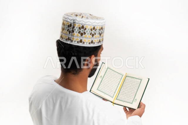 تلاوة وقراءة القرآن الكريم، بورتريه مقرب من الخلف لشاب عربي خليجي عماني يرتدي الدشداشة والكمه يحمل بيده المصحف الشريف، الإكثار من ذكر الله تعالى، الاجتهاد في العبادة، التقرب الى الله في شهر رمضان، الشعور بالخشوع والطمأنينة، خلفية بيضاء