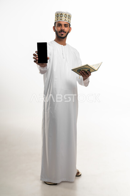 النظر الى الكاميرا بايماءات الخشوع والطمأنينة، بورتريه لشاب عربي خليجي عماني يرتدي الدشداشة والكمه يحمل بيده المصحف الشريف والهاتف المحمول، عرض شاشة سوداء فارغة على الموبايل، الاجتهاد في العبادة، التقرب الى الله بأداء الطاعات، خلفية بيضاء