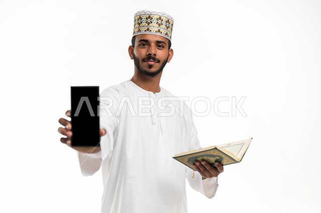 عرض شاشة سوداء فارغة على الموبايل، الاجتهاد في العبادة، النظر الى الكاميرا بايماءات الخشوع والطمأنينة، بورتريه مقرب لشاب عربي خليجي عماني يرتدي الدشداشة والكمه يحمل بيده المصحف الشريف والهاتف المحمول، التقرب الى الله بأداء الطاعات، خلفية بيضاء