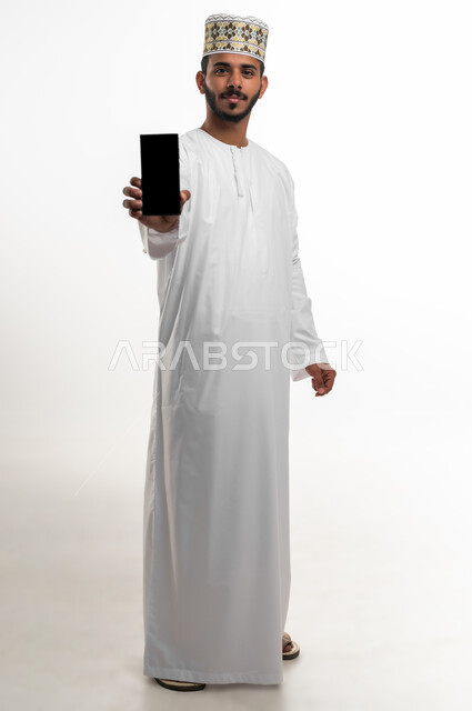 Using technology and advanced modern techniques, a portrait of a smiling Arab Gulf Omani young man wearing a dishdasha and cuff, displaying a blank black screen on a mobile phone, looking at the camera with an expression of pleasure, full-length body photo, white background.