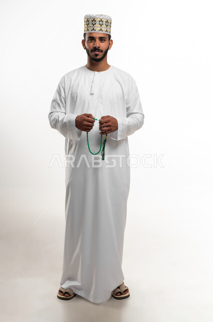 الإكثار من ذكر الله تعالى من خلال الإستغفار والدعاء, الإجتهاد في العباده والتقرب الى الله في شهر رمضان المبارك, مفهوم التسبيح والتهليل، بورتريه لشاب عربي خليجي عماني يرتدي الدشداشة والكمه ينظر الى الكاميرا يمسك بيده السبحه, الطول الكامل للجسم, خلفيه بيضاء