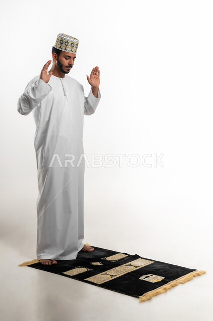 ايماءات التكبير لبدء الصلاة، أداء الفرائض بأوقاتها، بورتريه لشاب عربي خليجي عماني يرتدي الدشداشة والكمه يقف على السجادة، العبادة والتقرب إلى الله، الشعور بالخشوع والطمأنينة، الجسم بالطول الكامل، خلفية بيضاء