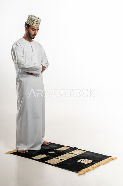 أهمية الالتزام بالصلاة على وقتها، الركن الثاني من أركان الإسلام، بورتريه لشاب عربي خليجي عماني يرتدي الدشداشة والكمه يقف على السجادة، التقرب من الله تعالى بأداء الفرائض والتزام أوامره, الشعور بالخشوع والطمأنينة، الجسم بالطول الكامل، خلفية بيضاء