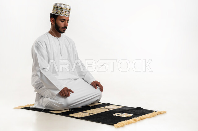 مفهوم أركان الاسلام والأيمان، الحفاظ على إقامة الصلوات الخمس بوقتها، التقرب الى الله تعالى بالإجتهاد في العبادة، بورتريه لشاب عربي خليجي عماني يرتدي الدشداشة والكمه يجلس على السجادة يقوم بأداء فريضة الصلاة، الشعور بالخشوع والطمأنينة، خلفية بيضاء