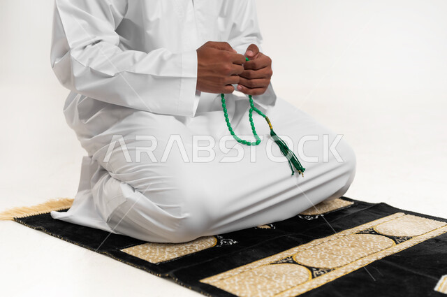 الإكثار من ذكر الله تعالى من خلال الإستغفار والدعاء, الإجتهاد في العباده والتقرب الى الله في شهر رمضان المبارك, مفهوم التسبيح والتهليل، بورتريه لشاب عربي خليجي عماني يرتدي الدشداشة والكمه يجلس على سجادة الصلاة يمسك بيده السبحه, خلفيه بيضاء