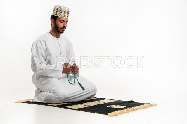مفهوم التسبيح والتهليل، الإجتهاد في العباده والتقرب الى الله في شهر رمضان المبارك, بورتريه لشاب عربي خليجي عماني يرتدي الدشداشة والكمه يجلس على سجادة الصلاة يمسك بيده السبحه, الإكثار من ذكر الله تعالى من خلال الإستغفار والدعاء, خلفيه بيضاء