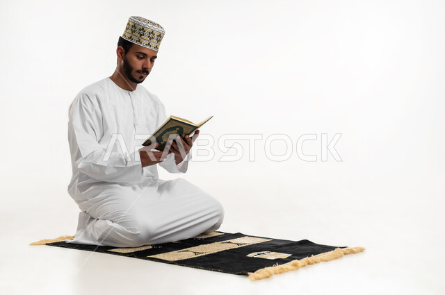 Striving in worship, getting closer to God in the month of Ramadan, remembering God often, reciting and reading the Holy Quran, portrait of a young Arab Gulf Omani man wearing a dishdasha and a turban sitting on a prayer rug and holding the Holy Quran in his hand, feeling reverence and tranquility, white background
