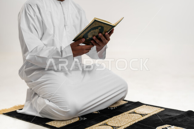 الشعور بالخشوع والطمأنينة، الاجتهاد في العبادة، التقرب الى الله في شهر رمضان، الإكثار من ذكر الله تعالى، تلاوة وقراءة القرآن الكريم، بورتريه  لشاب عربي خليجي عماني يرتدي الدشداشة والكمه يجلس على سجادة الصلاة ويحمل بيده المصحف الشريف، خلفية بيضاء