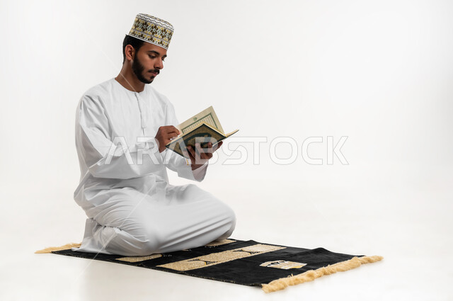 التقرب الى الله في شهر رمضان، الإكثار من ذكر الله تعالى، تلاوة وقراءة القرآن الكريم، الاجتهاد في العبادة، بورتريه  لشاب عربي خليجي عماني يرتدي الدشداشة والكمه يجلس على سجادة الصلاة ويحمل بيده المصحف الشريف،  الشعور بالخشوع والطمأنينة، خلفية بيضاء