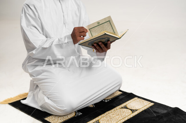 الاجتهاد في العبادة، التقرب الى الله في شهر رمضان، الإكثار من ذكر الله تعالى، تلاوة وقراءة القرآن الكريم، بورتريه  لشاب عربي خليجي عماني يرتدي الدشداشة والكمه يجلس على سجادة الصلاة ويحمل بيده المصحف الشريف،  الشعور بالخشوع والطمأنينة، خلفية بيضاء
