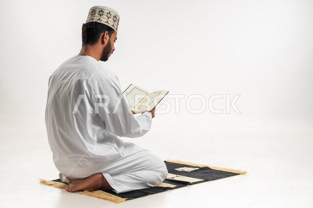 الإكثار من ذكر الله تعالى، تلاوة وقراءة القرآن الكريم، بورتريه من الخلف لشاب عربي خليجي عماني يرتدي الدشداشة والكمه يجلس على سجادة الصلاة ويحمل بيده المصحف الشريف، الاجتهاد في العبادة، التقرب الى الله في شهر رمضان، الشعور بالخشوع والطمأنينة، خلفية بيضاء