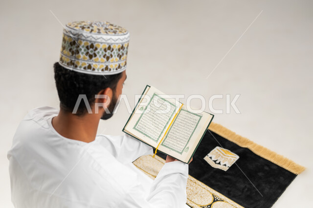 تلاوة وقراءة القرآن الكريم، بورتريه مقرب من الخلف لشاب عربي خليجي عماني يرتدي الدشداشة والكمه يجلس على سجادة الصلاة ويحمل بيده المصحف الشريف، الإكثار من ذكر الله تعالى، الاجتهاد في العبادة، التقرب الى الله في شهر رمضان، الشعور بالخشوع والطمأنينة، خلفية بيضاء
