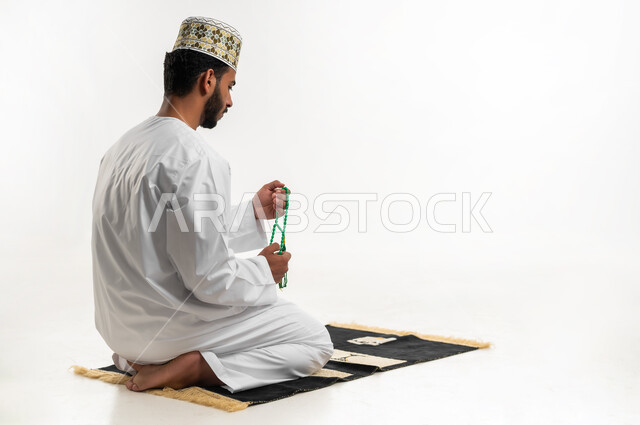 الإجتهاد في العباده والتقرب الى الله في شهر رمضان المبارك, مفهوم التسبيح والتهليل، بورتريه لشاب عربي خليجي عماني يرتدي الدشداشة والكمه يجلس على سجادة الصلاة يمسك بيده السبحه, الإكثار من ذكر الله تعالى من خلال الإستغفار والدعاء, خلفيه بيضاء