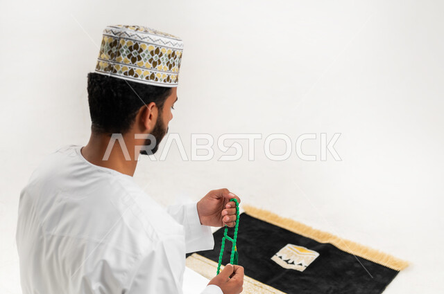 مفهوم التسبيح والتهليل، الإجتهاد في العباده والتقرب الى الله في شهر رمضان المبارك,بورتريه لشاب عربي خليجي عماني يرتدي الدشداشة والكمه يجلس على سجادة الصلاة يمسك بيده السبحه, الإكثار من ذكر الله تعالى من خلال الإستغفار والدعاء, خلفيه بيضاء